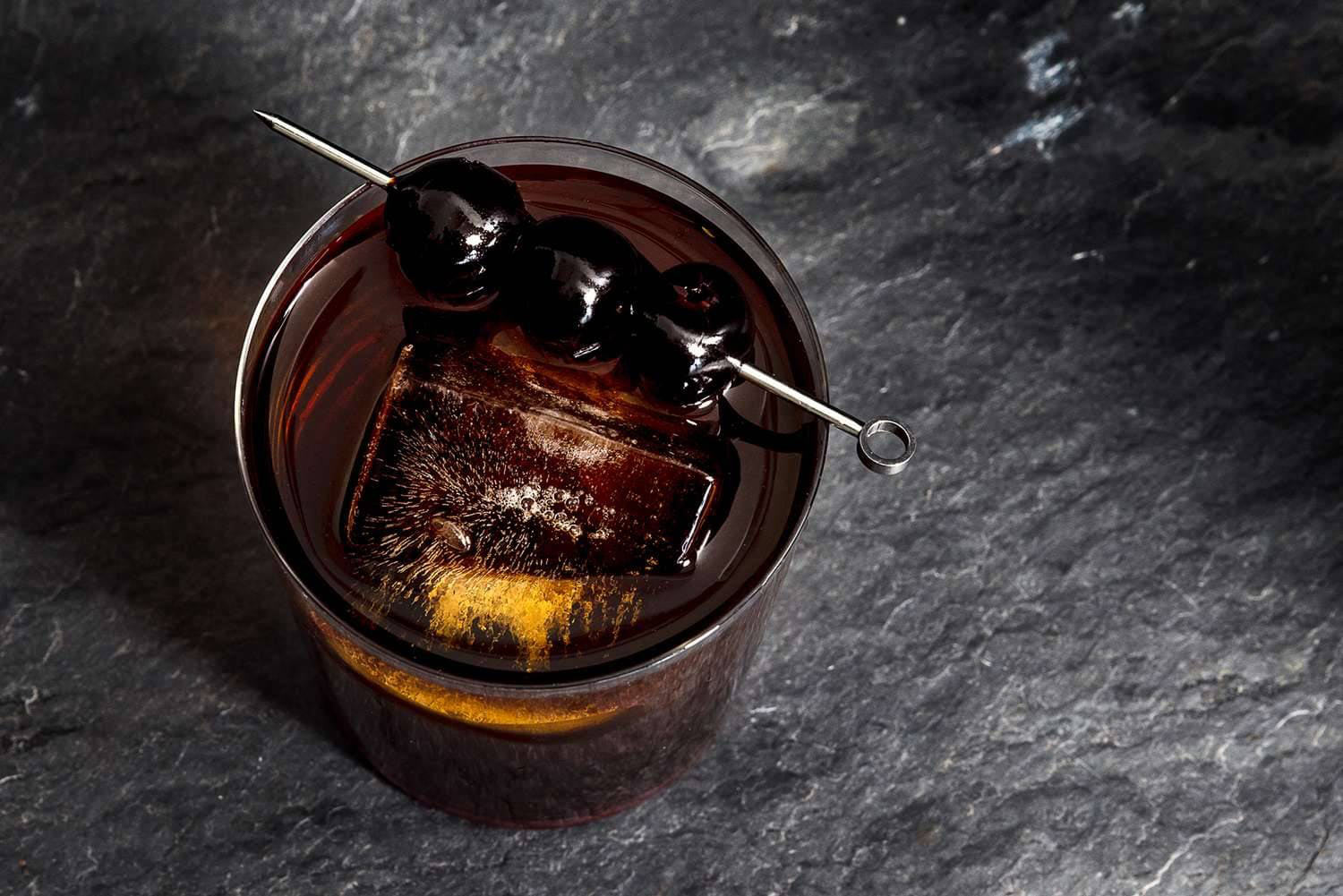 Dark cocktail with a large ice cube and three cherries on a skewer